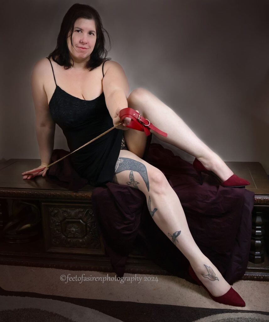 Photograph of a dominatrix in a black dress sitting on a brown wooden bench, one leg popped up on the bench, leaning off onto one arm, holding a red leather collar and gold chain, smiling at the camera.