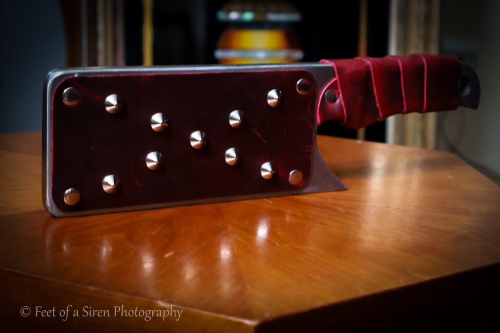 Steel cleaver shaped BDSM paddle. Red leather wrapped handle and leather stacked riveted face.