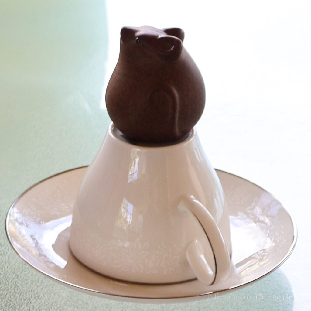 Small dormouse figurine sitting atop an overturned teacup on a saucer on a table.