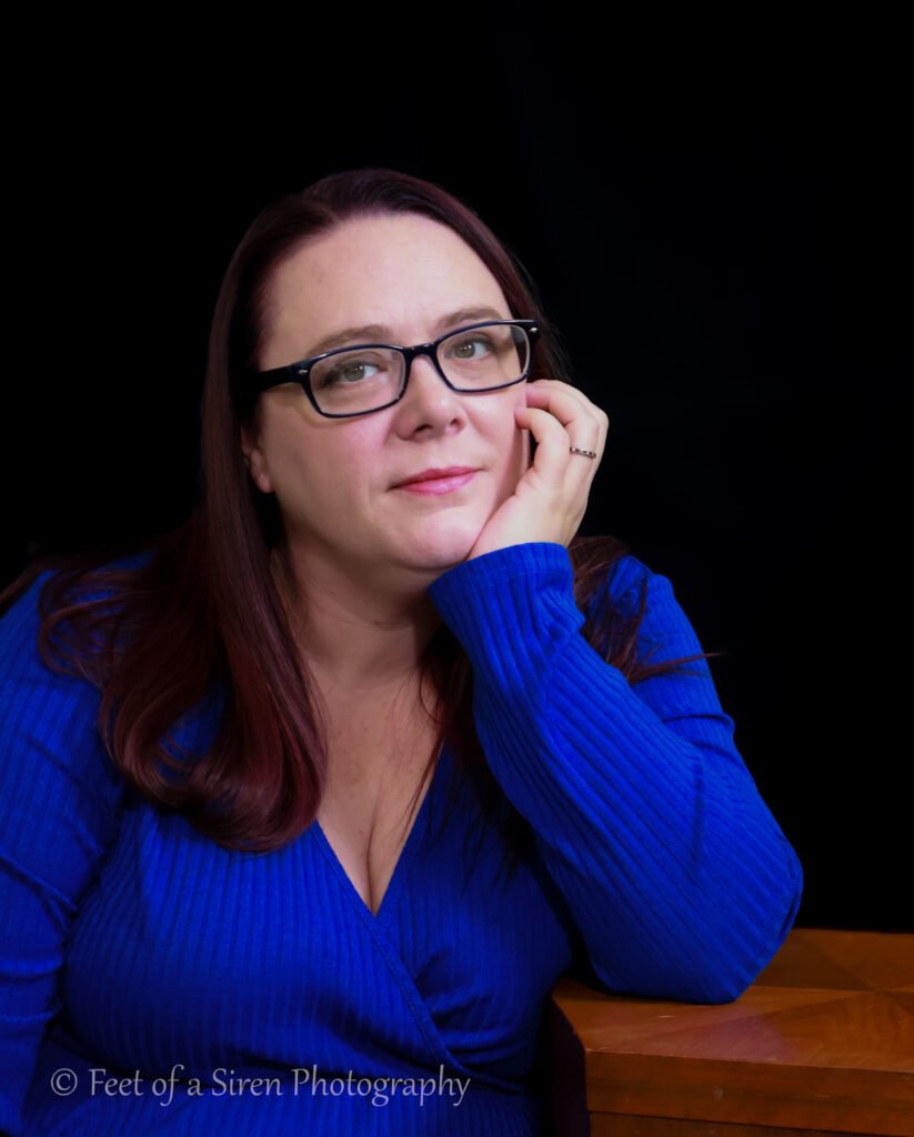 Woman in blue long sleeve dress, elbow on a wooden table, hand under her chin, smiling.