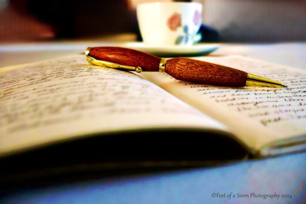 Open Notebook with writing on the pages, a pen across the open face, a teacup and saucer in the background.