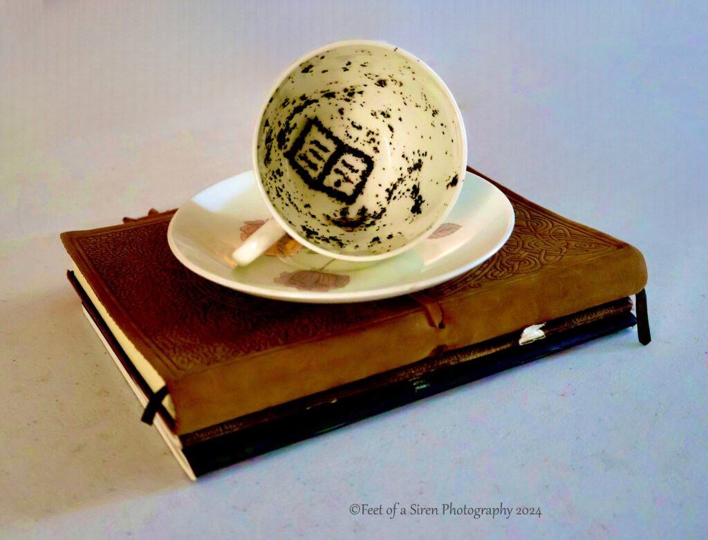 Two notebooks stacked, with a cup and saucer on top. Remnant tea leaves in the shape of a book inside the cup.