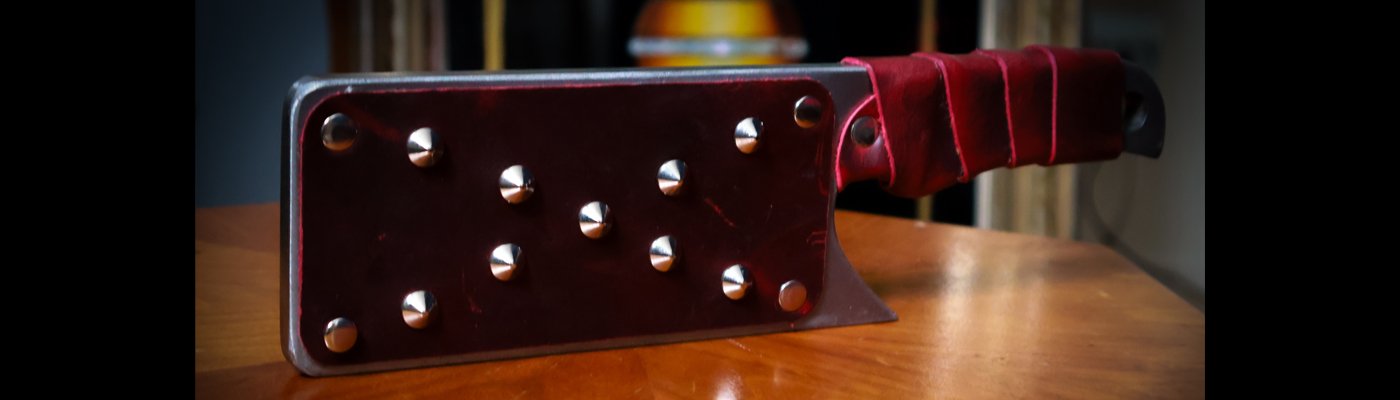 Color photograph of a steel paddle in the shape of a cleaver, with red leather wrapped handle, and red leather surfaced face, covered in silver rivets.