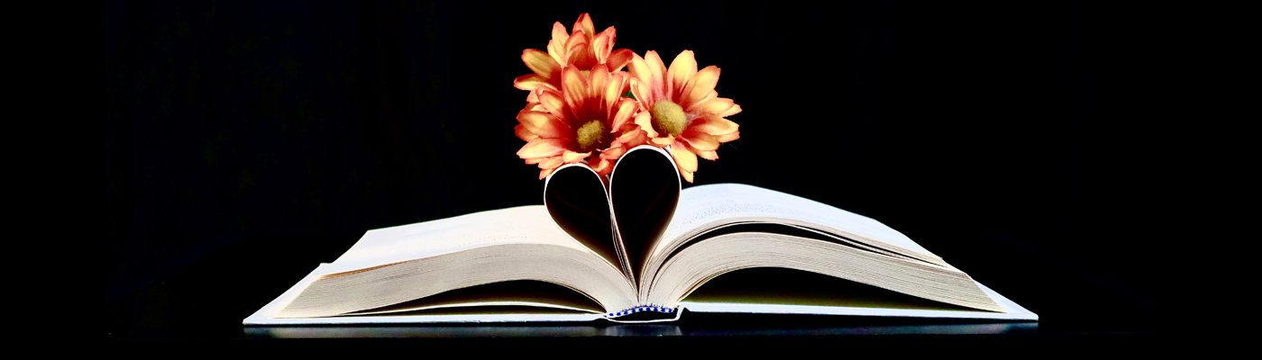 Color photograph of an open faced book with some pages curled in to form a heart. Yellow and red flowers sitting atop the heart.