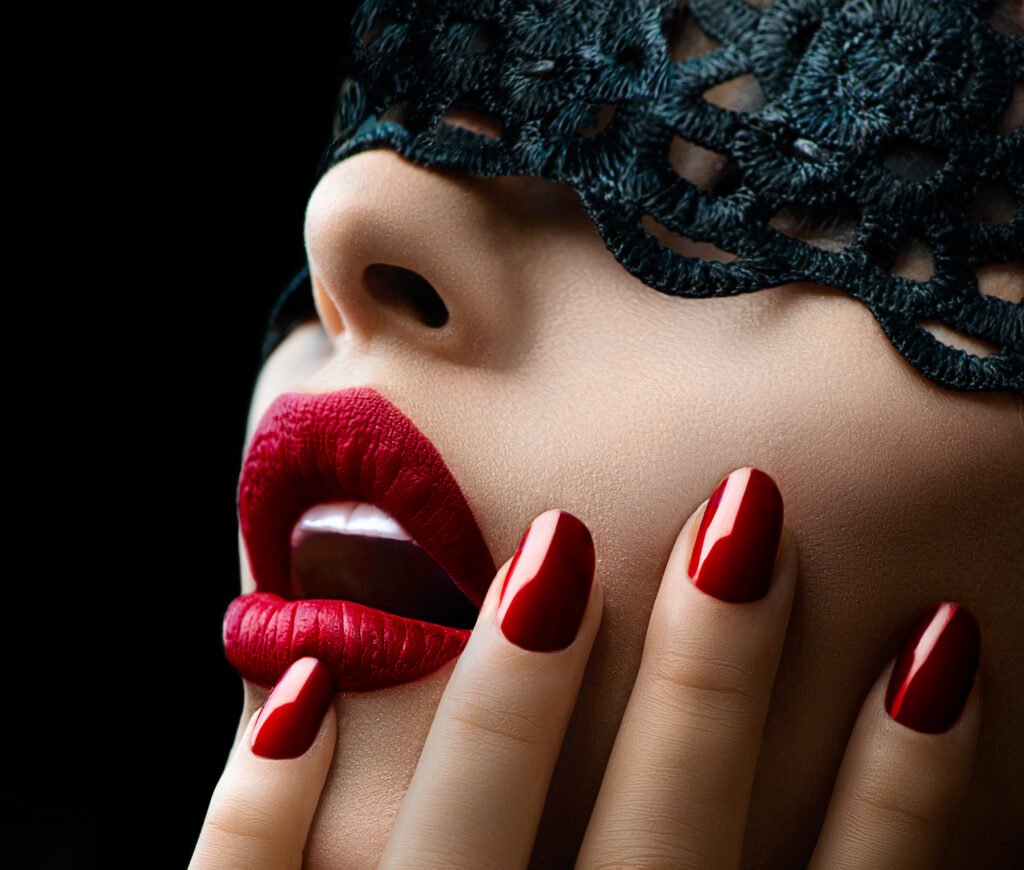 photo of a woman with red nail polish and lipstick, touching her face, a black lace mask covering her eyes.