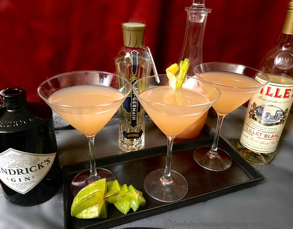 Three martini glasses filled with a peach colored liquid, surrounded by three alcohol bottles, with star fruit at their bases sliced into a few pieces. Red backdrop, grey tablecloth.